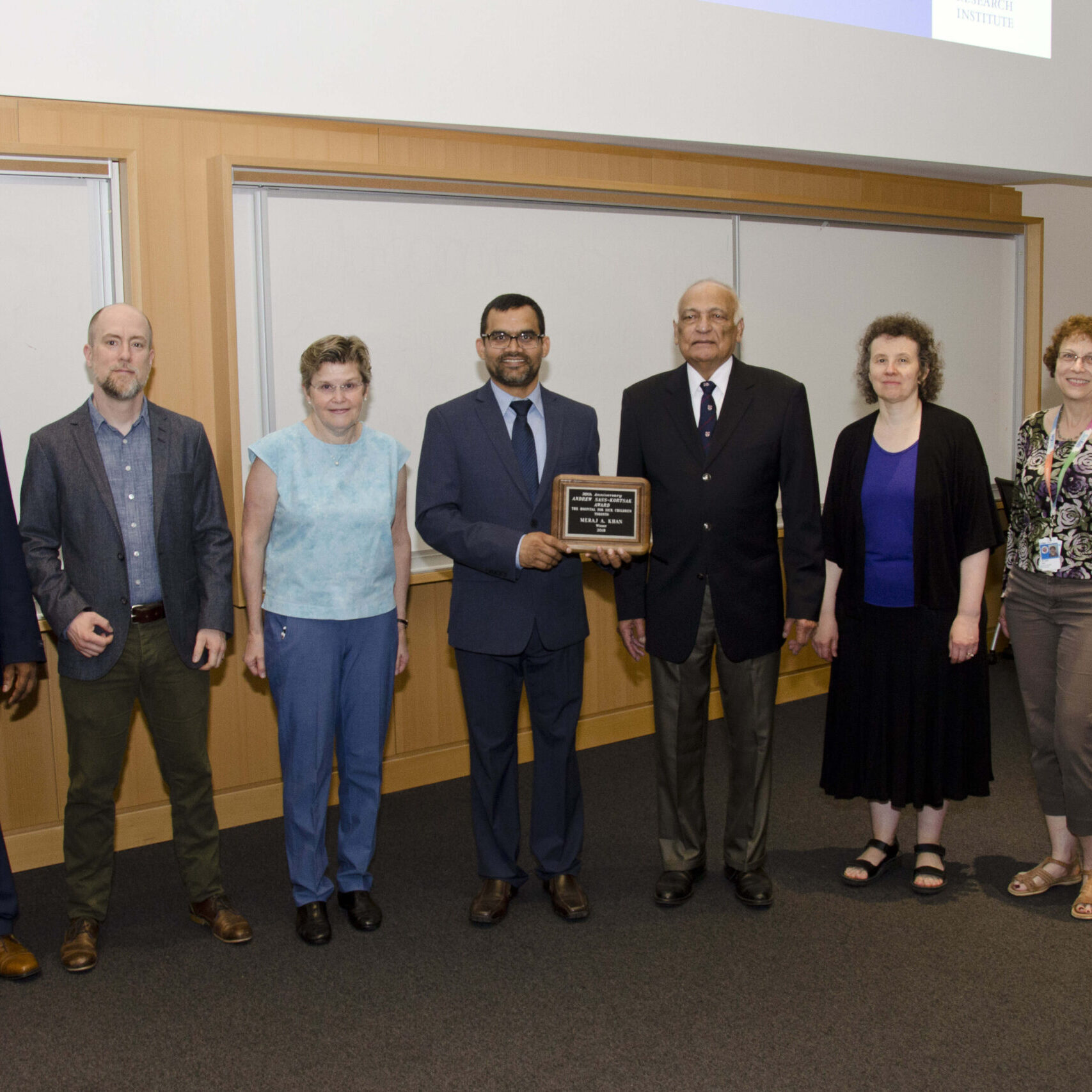 Meraj A Khan in the center, holding the Sass-Kortsak Award, a prestigious SickKids Award. He is surrounded by 7 other people to his left and right, who are part of the award committee.
