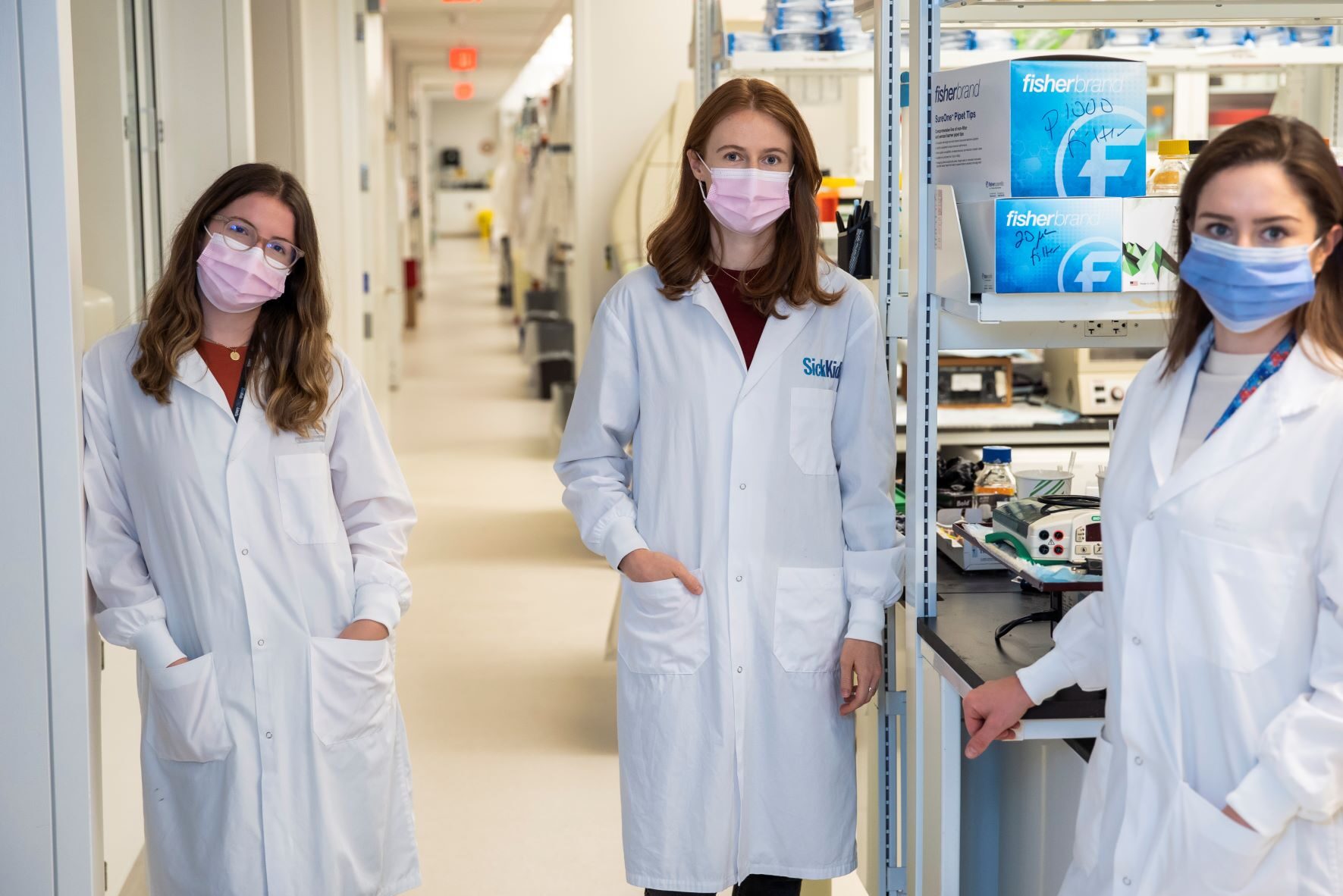 3 women in lab coats, masked, in a lab