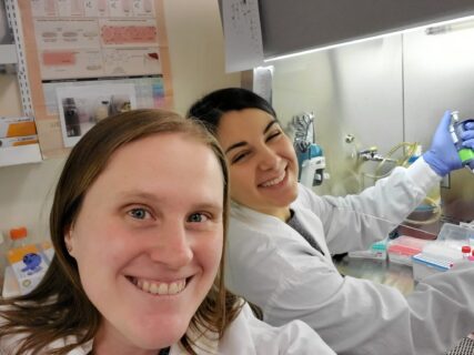 Dr. Victoria Forster and Dr. Melissa Galati posing in front of a biosafety cabinet at the Hospital for Sick Children. Dr. Melissa Galati is holding a pipette.