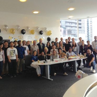 A large group of lab members standing together smiling, surrounded by balloons, cakes and a happy birthday banner