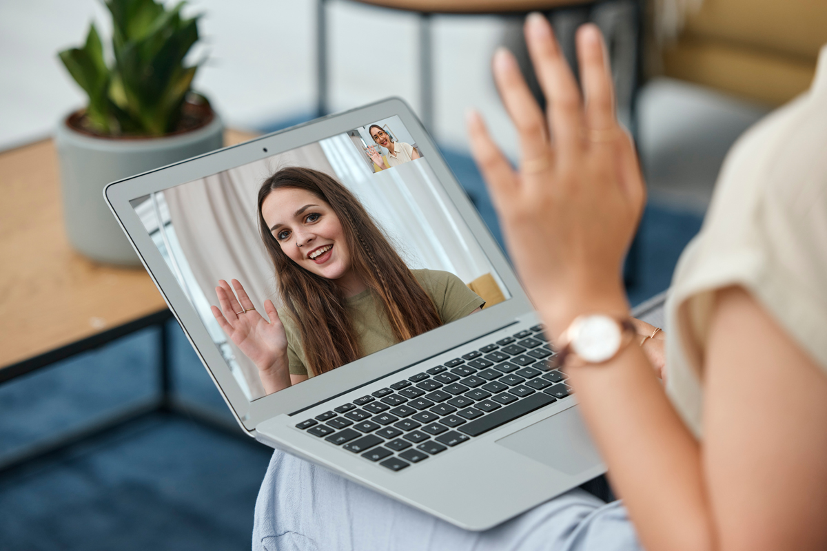 A person on a video call on their laptop and waving. The laptop screen shows a woman who is waving as well.