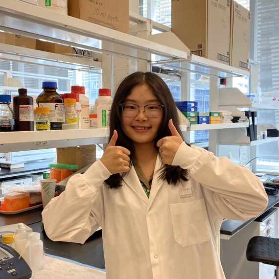 Portrait of Xi in the wet lab, giving a double thumbs up to the camera. She is wearing a white lab coat, rectangle-rimmed glasses, and smiling at the camera.