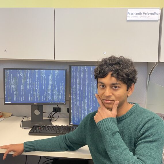 Picture of Prashanth sitting at his computer desk with a serious "thinking" look on his face and hand on his chin. The monitors behind him display 0s and 1s.