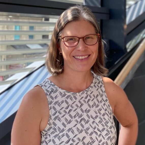 Portrait of Lisa. She is standing on a staircase, in front of windows, wearing a white and grey tank top, red, square-rimmed glasses, and smiling at the camera.