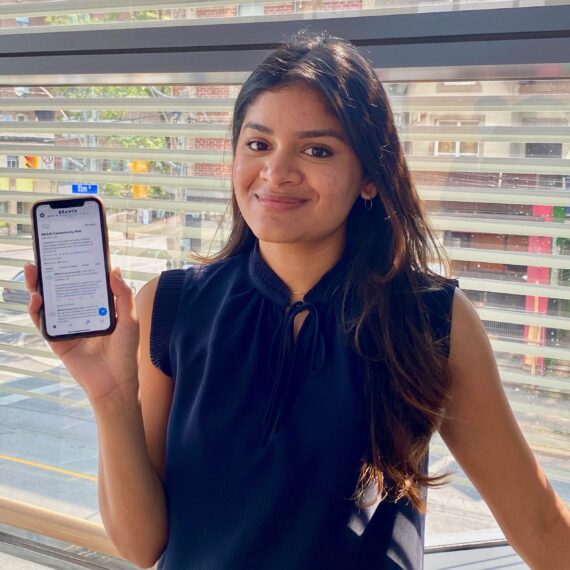 Portrait of Eman standing in front of a window, holding her cellphone which shows the lab's twitter page.