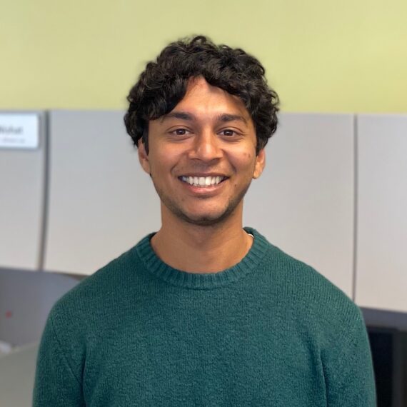 Portrait of Prashanth standing in the lab. He is wearing a green sweater and smiling at the camera.