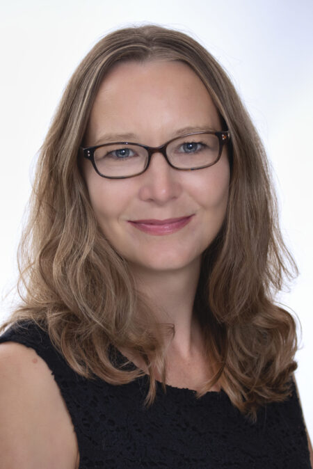 Headshot of Anne. She is wearing a black blouse, rectangular rimmed glasses, and smiling at the camera.