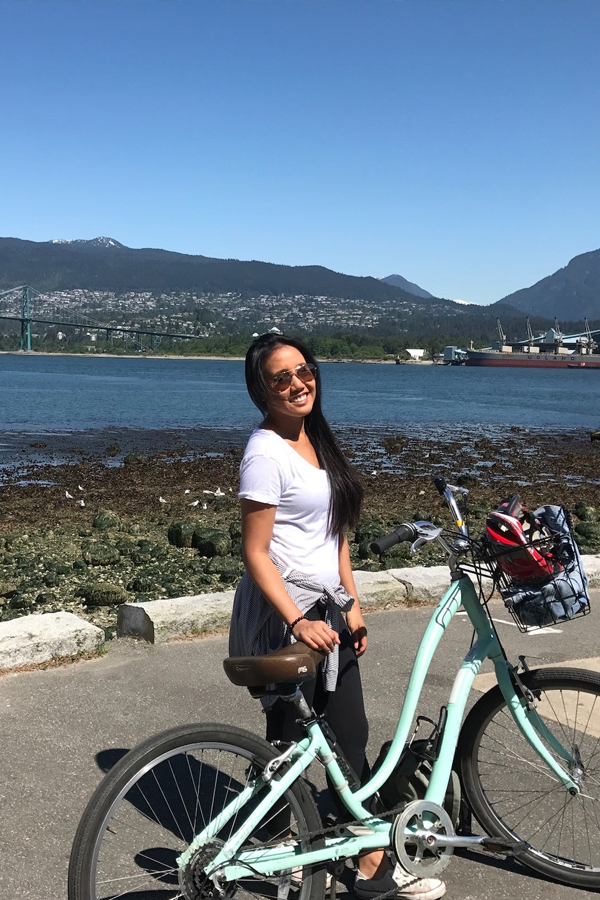 Photo of Alison Cateyano standing next to a bicycle in the foreground with a scenic view of water, a bridge and mountains in the background.