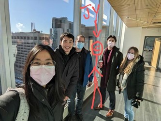 lab_team_pic Five people standing in a hallway with large windows overlooking a cityscape. Four of them are wearing masks. A funny stick figure has been edited on the picture to represent Dr. Ji-Young Youn.