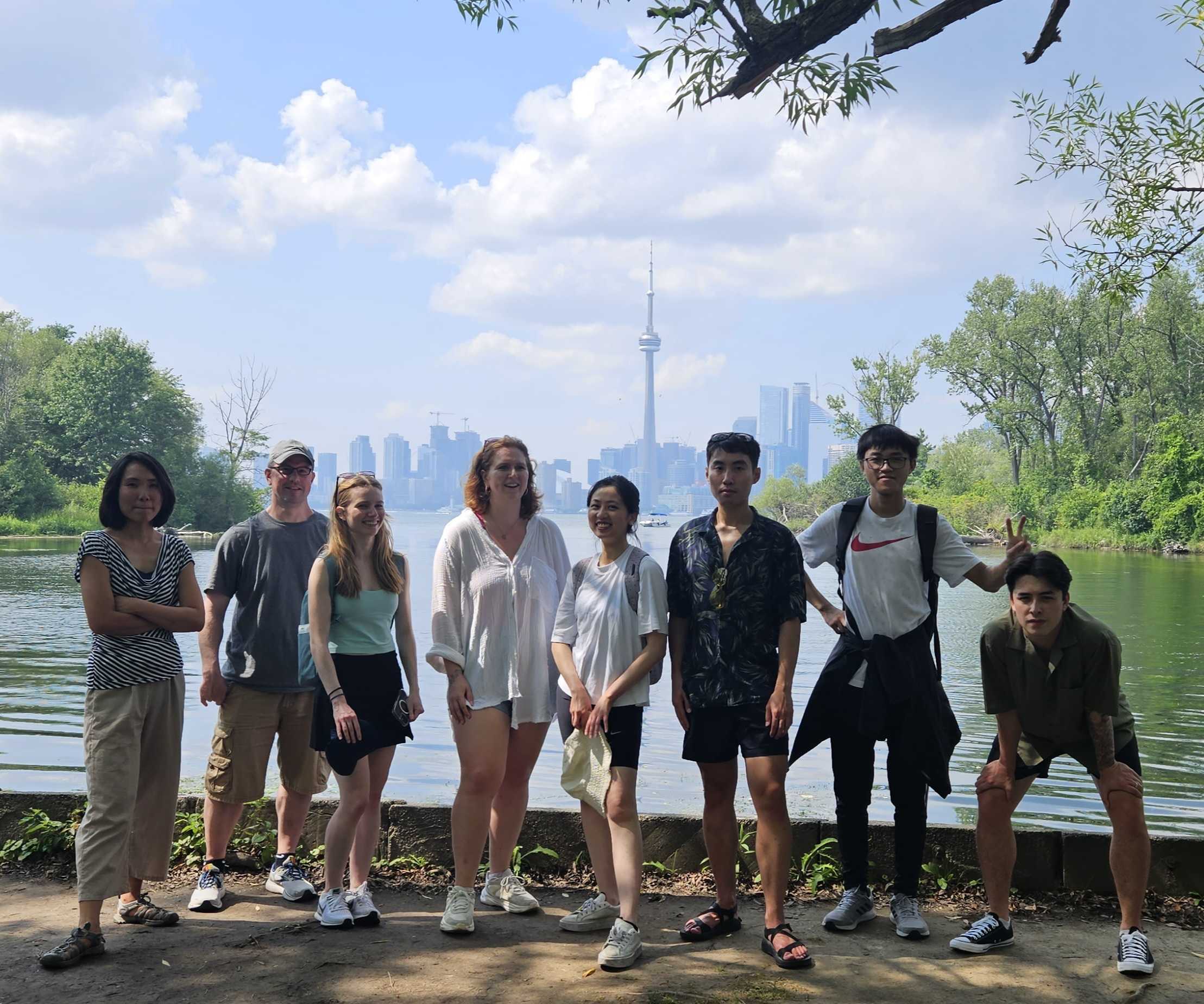 20230901_233345 Eight people posing for a group photo outdoors near a body of water, with the Toronto skyline and CN Tower visible in the background.