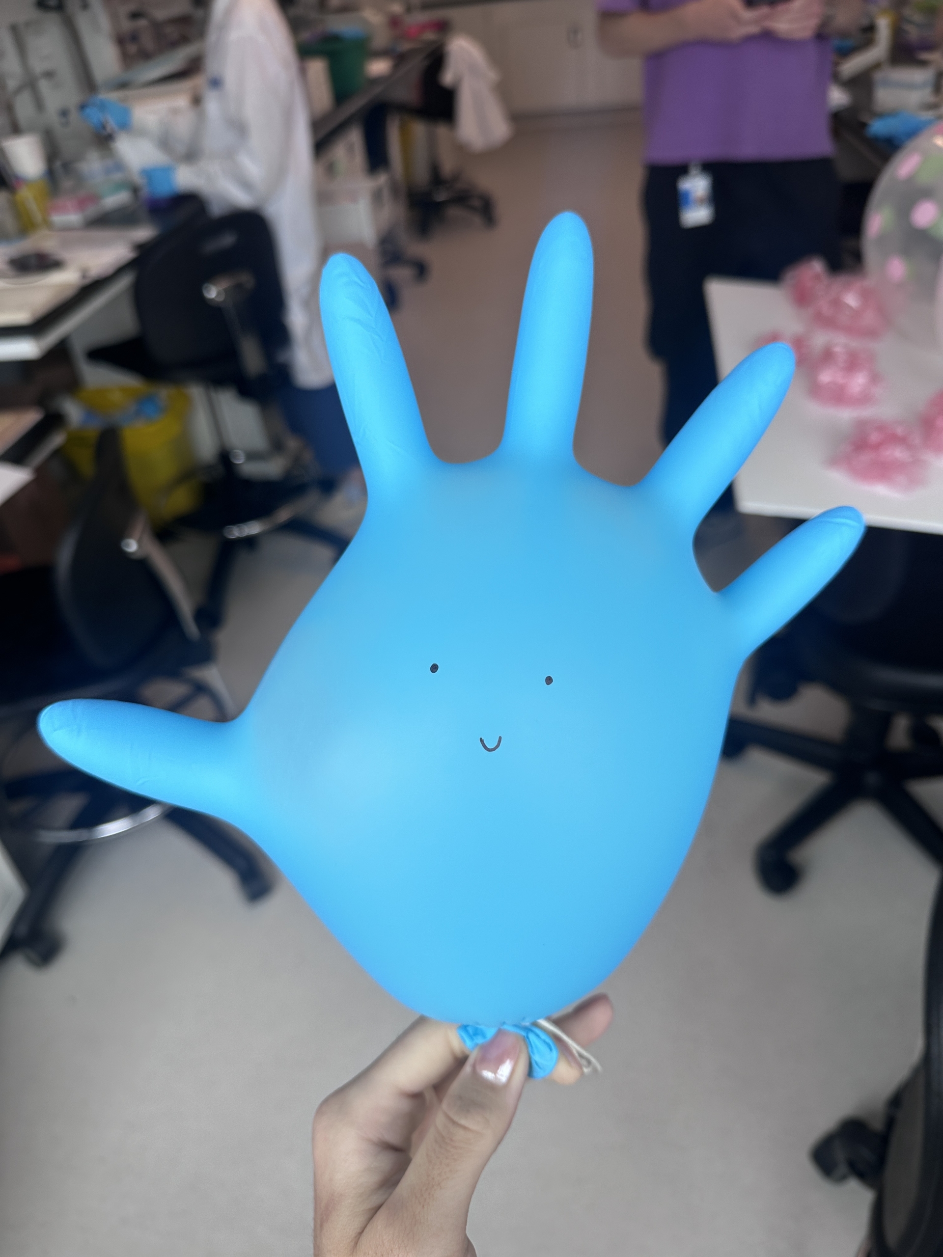 IMG_2869 Inflated blue latex glove with a smiley face drawn on it; background shows a lab or medical setting with equipment, chairs, and people in lab coats.