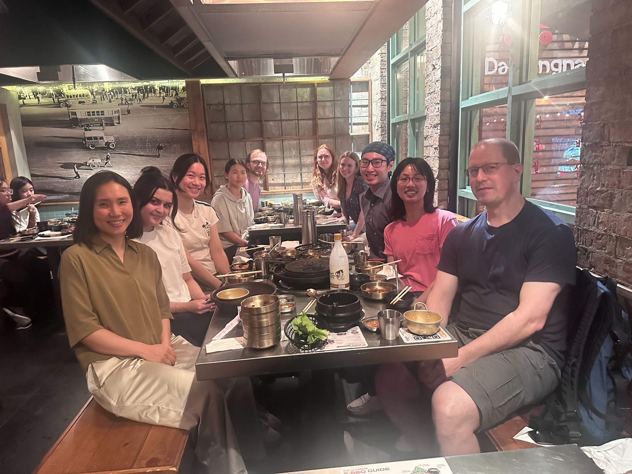 IMG_2873 Group of people seated around a table in a restaurant with dishes, bowls, and utensils; background features brick walls and green-framed windows.