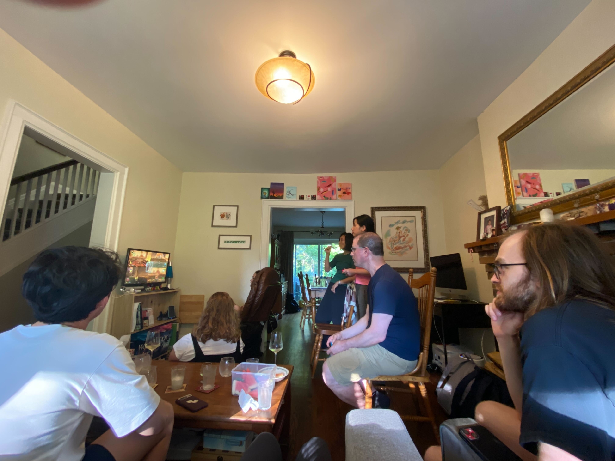 IMG_6298 Group of people gathered in a living room with a ceiling light, staircase, furniture, and a TV on the left; framed pictures and artwork decorate the walls.