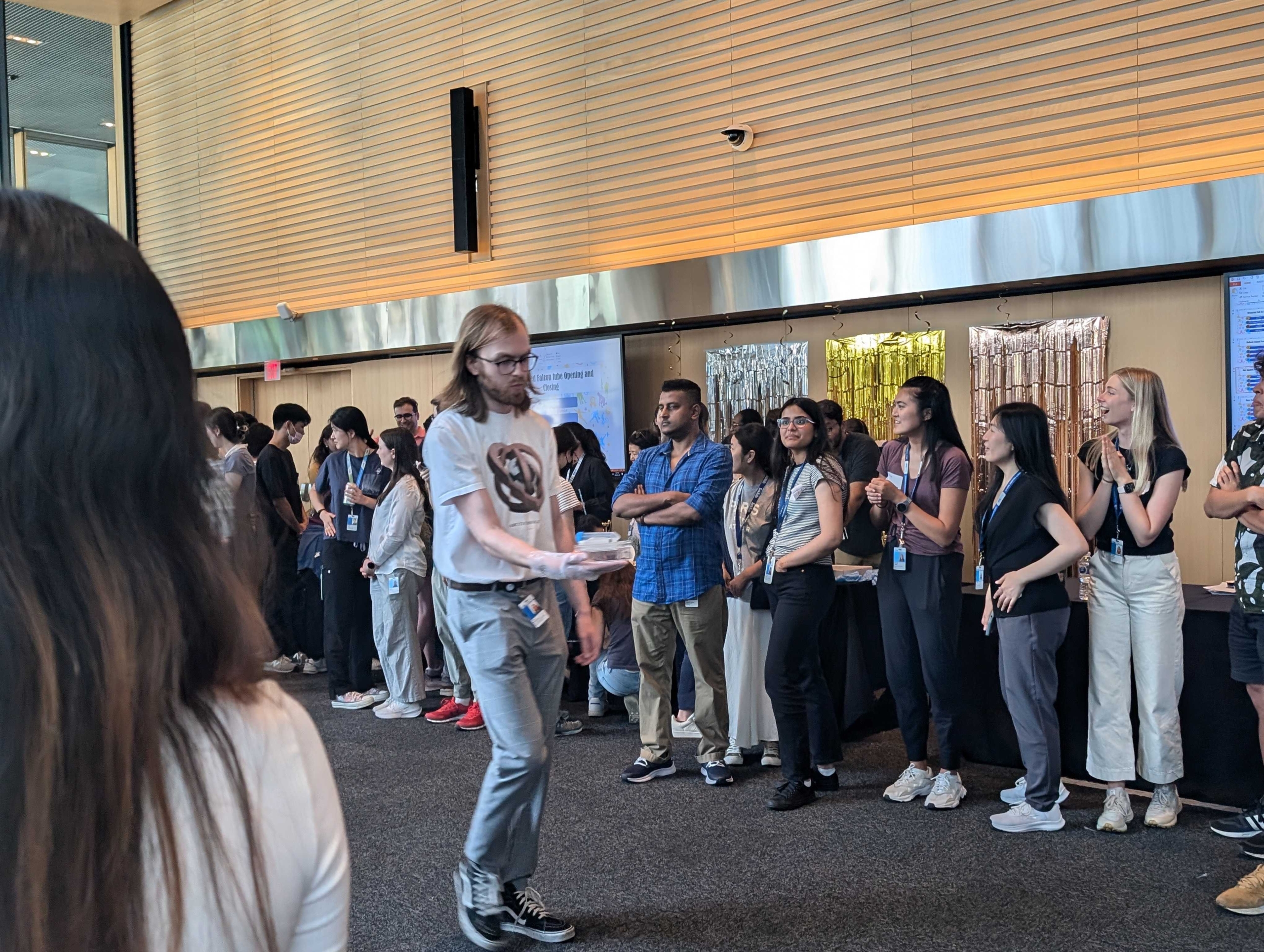 Group of people gathered indoors near wooden panel walls and large windows; one person wearing glasses walks toward the camera holding a tray.