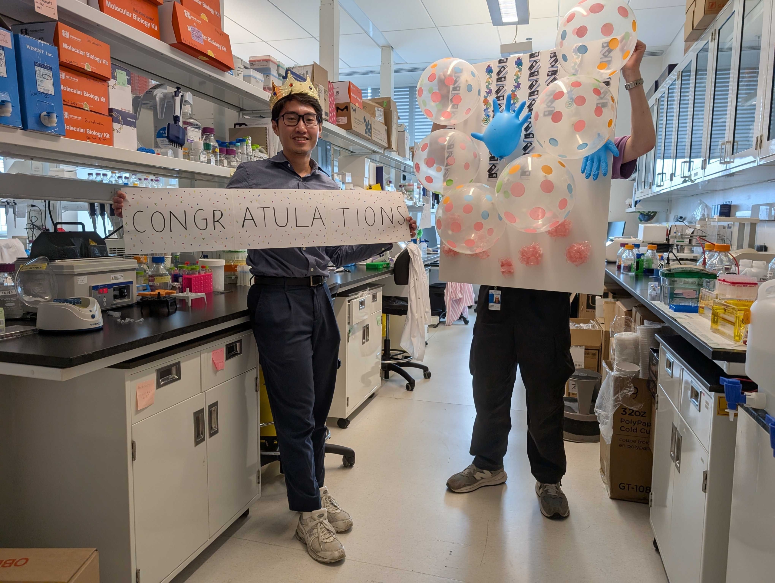 Two people in a lab; one holds a “CONGRATULATIONS” sign, the other holds a board with balloons, including pink and white polka-dotted ones. Lab equipment and cabinets are visible in the background.
