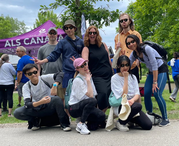 Group of people posing with peace signs and sunglasses, outdoors in front of a purple tent labeled “ALS CANADA,” with trees and other individuals visible in the background.