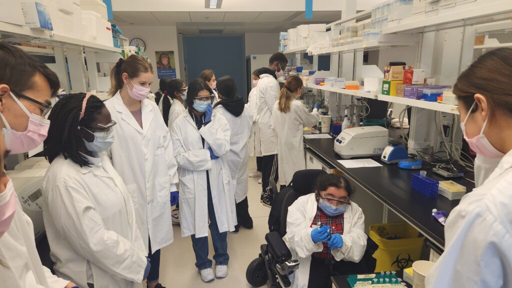 Group of students in lab coats standing in lab watching someone using a pipet